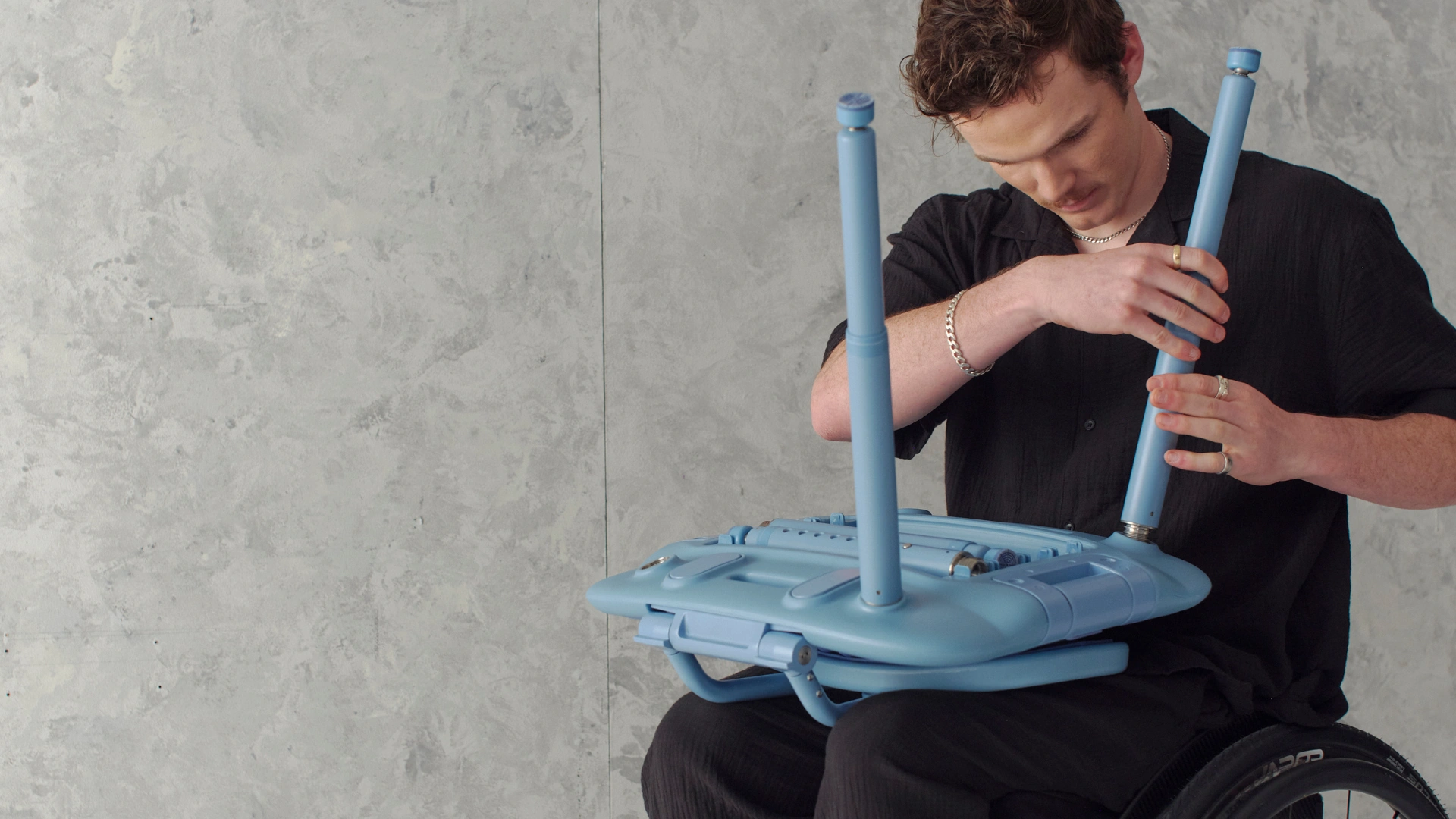 (Fletch) Man with light brown hair is sitting in his wheelchair. He is assembling a blue oasis chair on his lap, screwing in one of the telescopic legs.