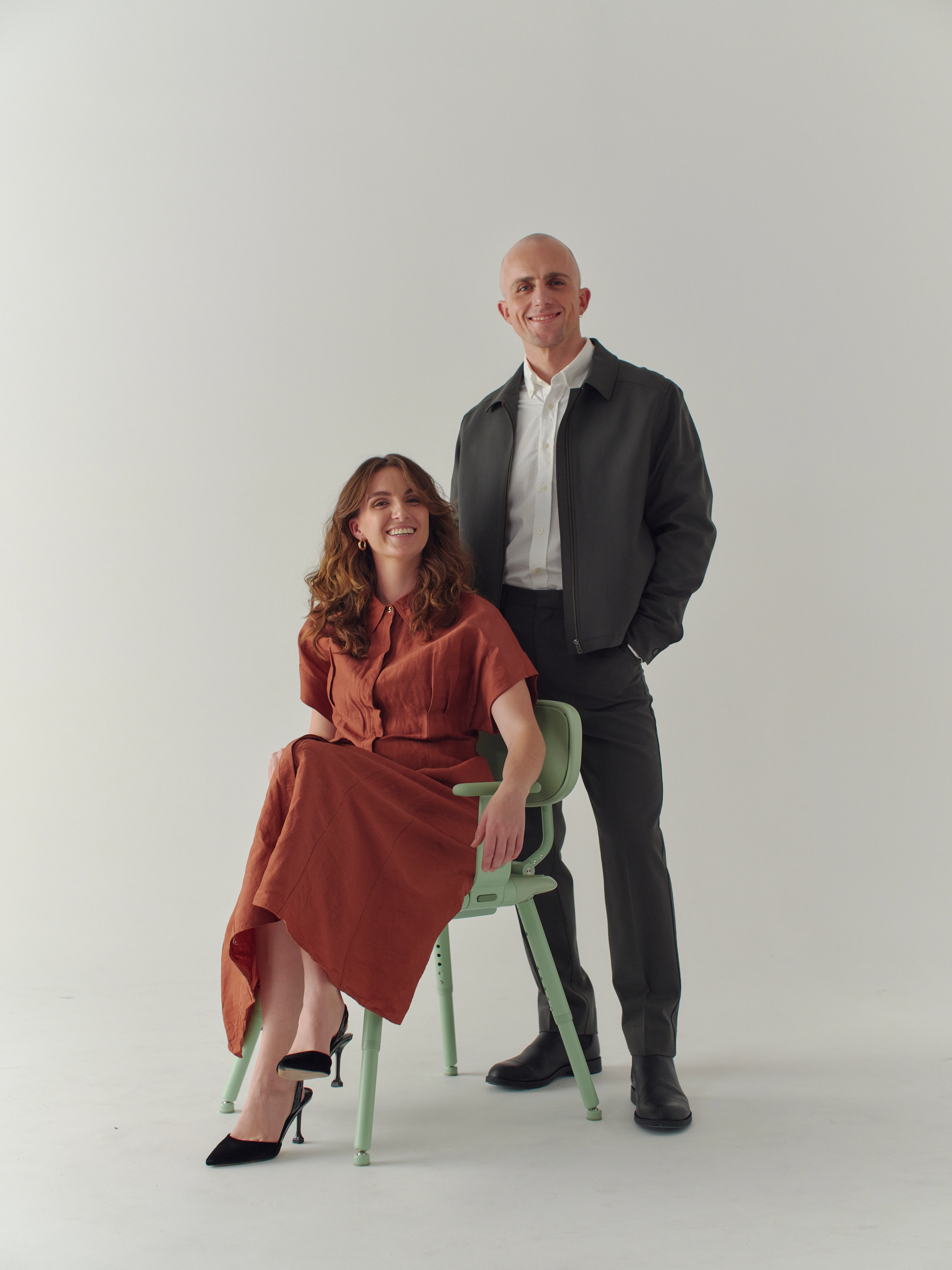 Woman in orange dress siits on green shower chair. Man strands behind her green pants and jacket