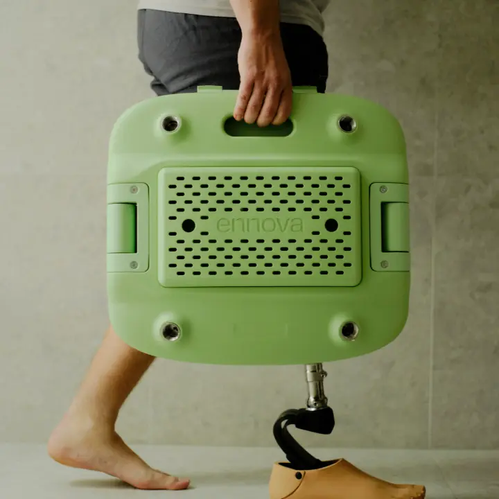 A person with a prosthetic leg is carrying a packed down, transportable pastel green Oasis shower chair with one hand.