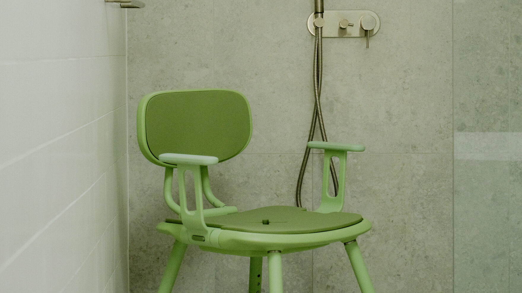 A pastel green Oasis shower chair is positioned in a tiled shower. The foldable armrests are up and ready.