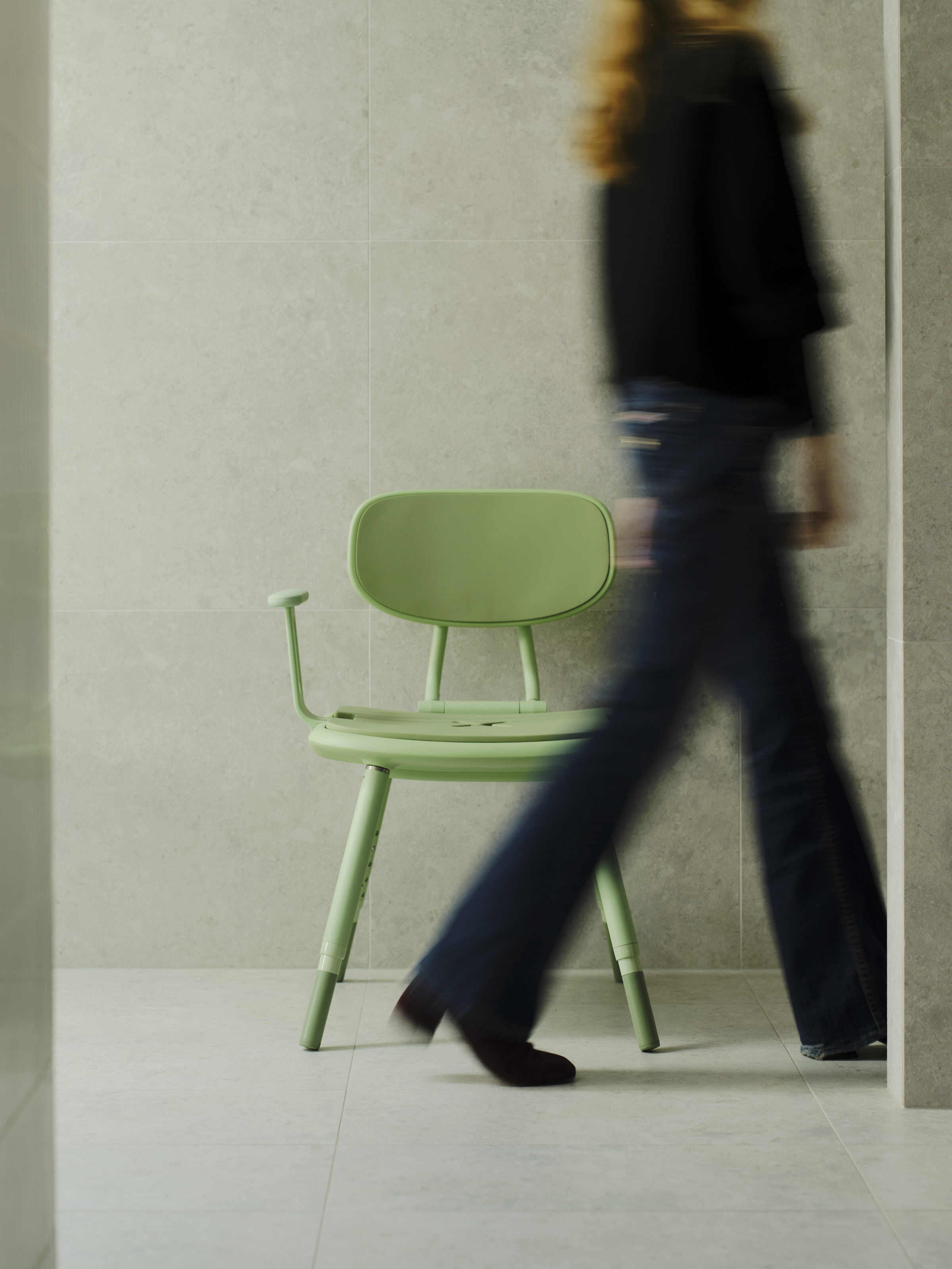 An image of a women in a black top and jeans is walking past the Oasis chair. A pastel green Oasis shower chair is positioned and ready in a tiled shower with the armrests up. The height adjustable legs are at their lowest setting.