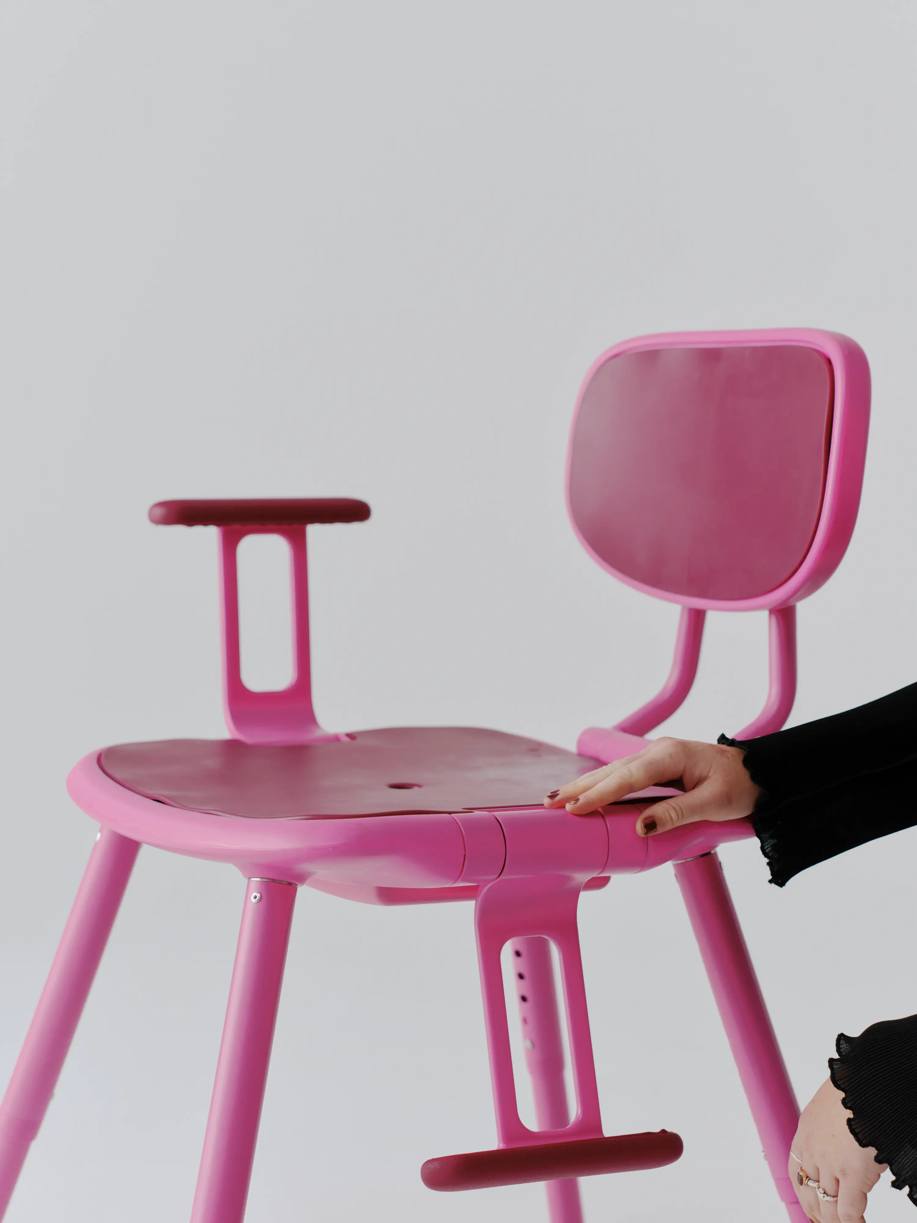 Pink height-adjustable shower chair with a padded seat, backrest and armrests, shown against a light grey background while a person adjusts the chair.