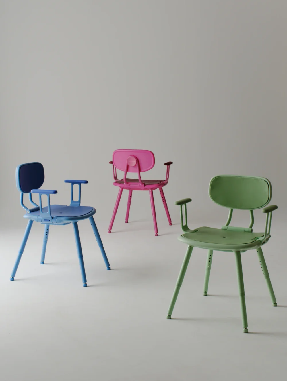 Three height-adjustable shower chairs arranged in a minimalist studio setting. A blue chair sits on the left, a pink chair is positioned behind it in the centre, and a green chair is in the foreground on the right. Each chair features a padded backrest, bi-directional armrests, contoured seat and four adjustable legs.