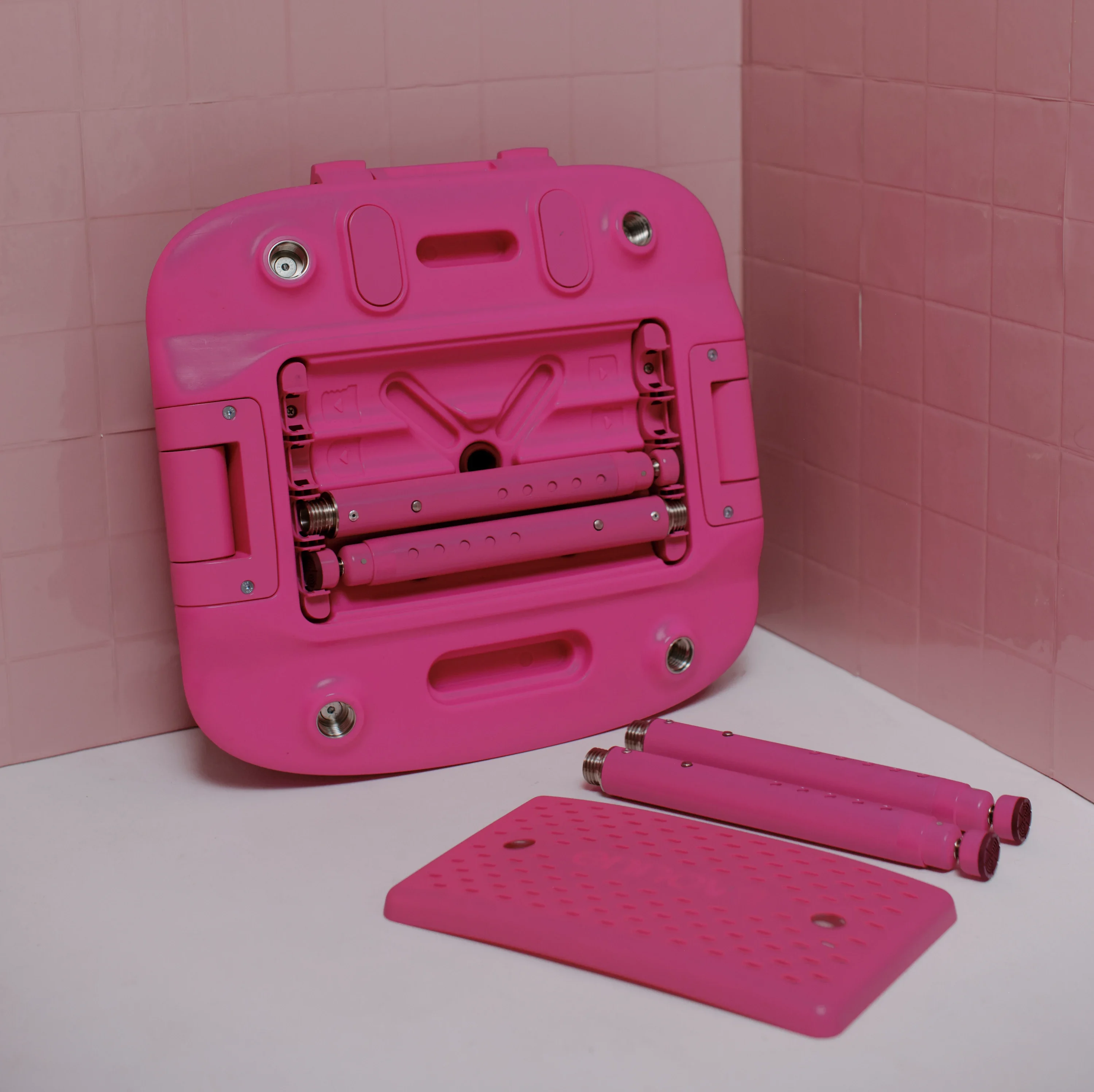 Pink Oasis Chair in its packed down form. Two adjustable legs and the leg storage cover are on the floor, with the oasis chair resting against a pink wall